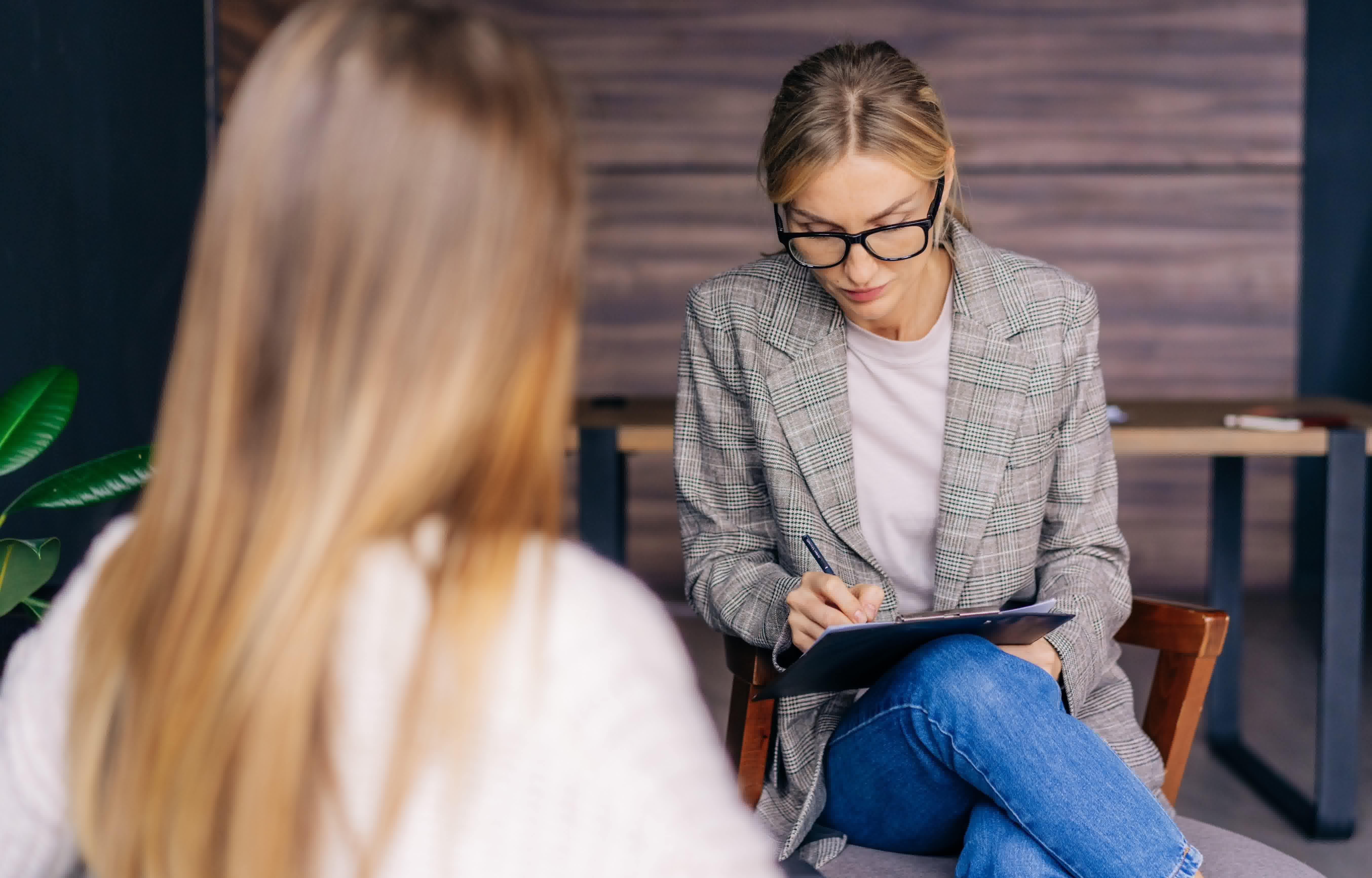 Individuelles Coaching im Pflegebereich: Eine professionelle Beraterin mit Brille macht sich während eines vertraulichen Gesprächs konzentriert Notizen auf einem Klemmbrett.