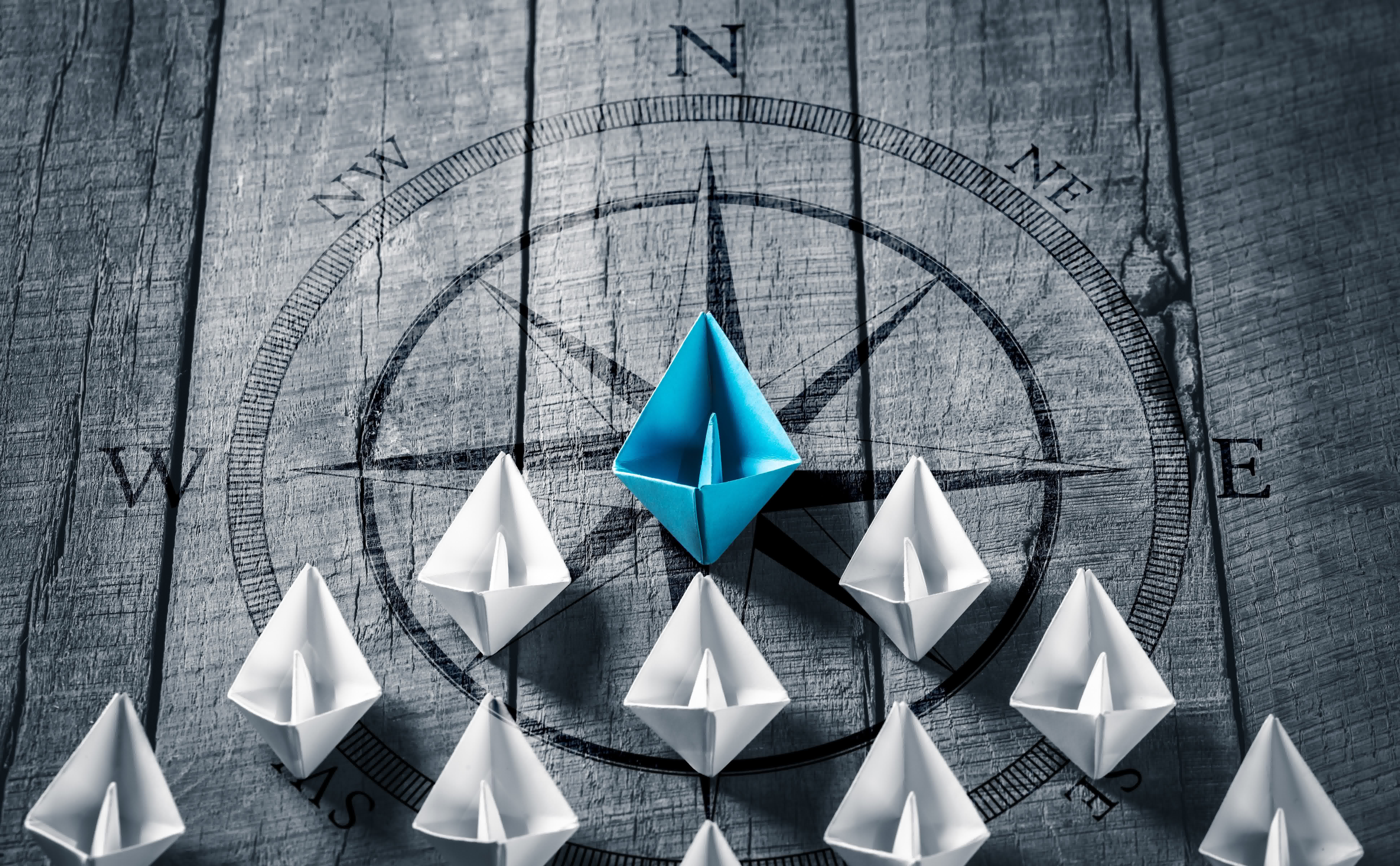 Metapher für Beratung, Weiterbildung und Forschung in der Pflege: Ein blaues Papierboot führt eine Gruppe weißer Boote auf einem hölzernen Untergrund mit Kompass im Hintergrund an.